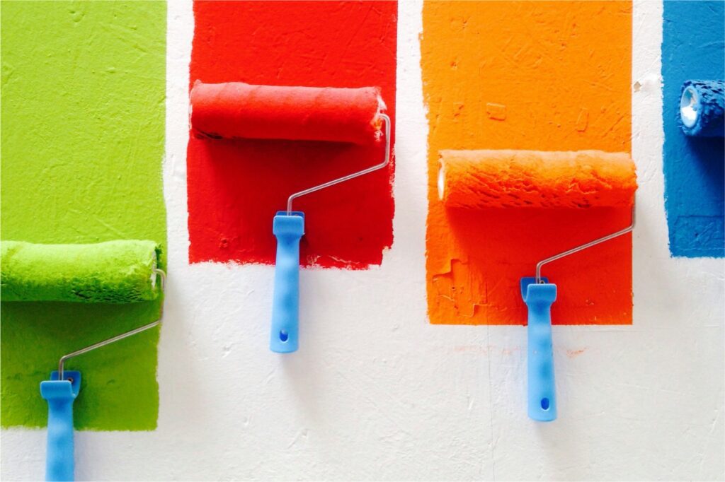 Rodillos de pintura aplicando diferentes colores vibrantes sobre una pared blanca, representando opciones de colores para pintar una casa.